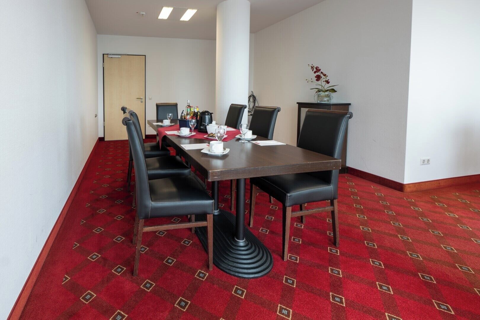 Un salón de conferencias con piso alfombrado en rojo, una mesa de madera, sillas negras y un jarrón con flores sobre una mesita auxiliar.