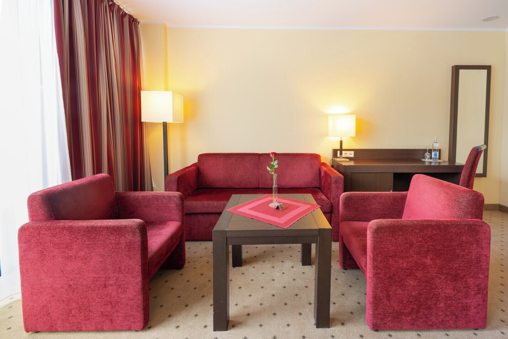 A hotel room with a red sofa, two red armchairs, a small table with a red placemat, and two bedside lamps.