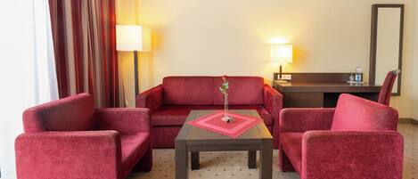 Habitación de hotel con sofá rojo, dos sillones rojos, una mesita con posavasos rojo y dos lámparas de noche.