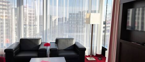Een moderne hotelkamer met een groot raam, een zwarte leren bank, een klein tafeltje met een bloemenvaas, en een flatscreen televisie.