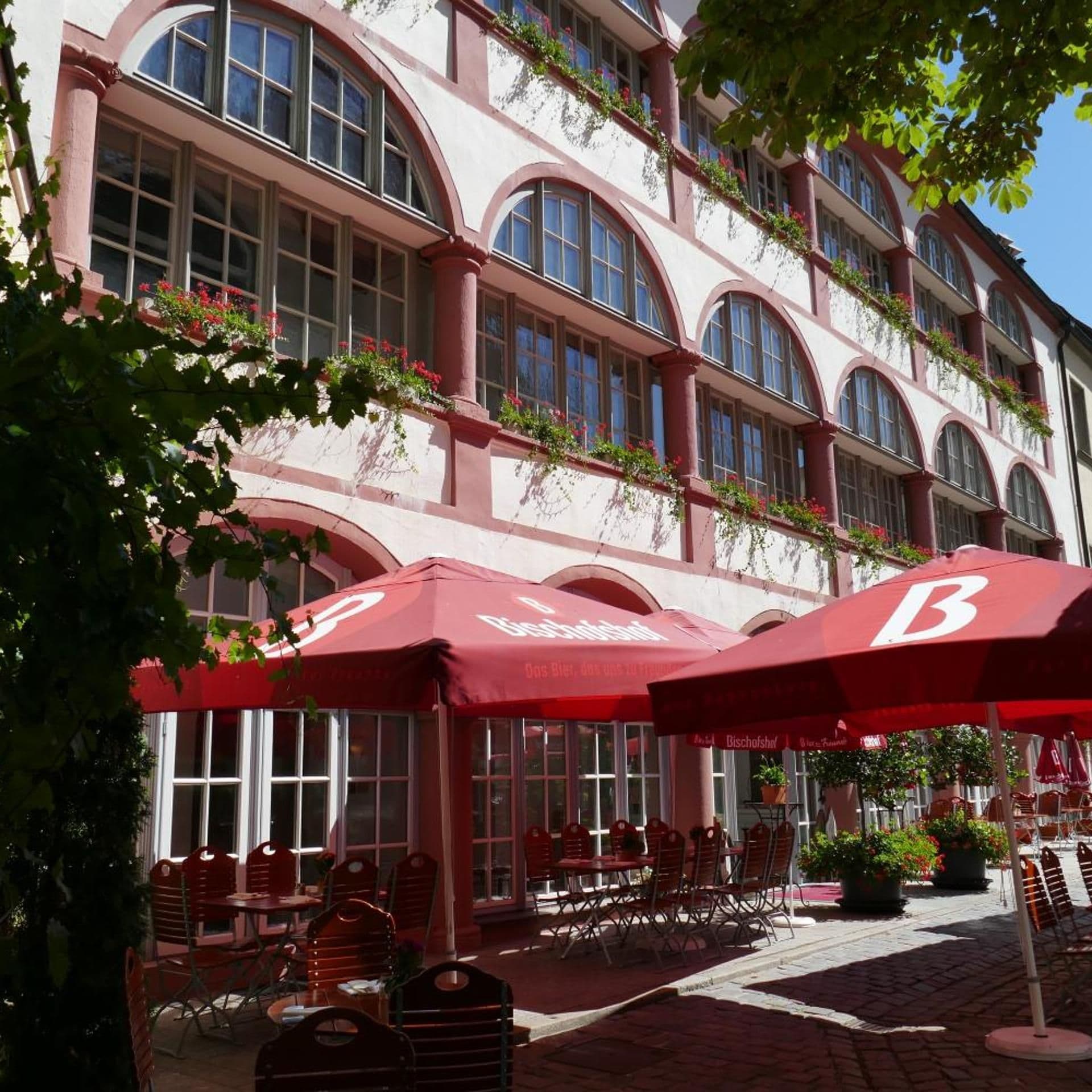 Un edificio con ventanas de arco y toldos rojos, que cuenta con asientos al aire libre y plantas en macetas.