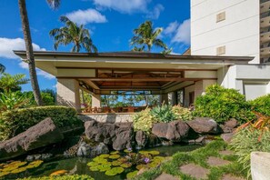 Um hotel moderno com área de estar coberta ao ar livre, um pequeno lago com nenúfares e paisagismo tropical.