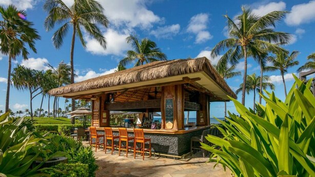 A thatched-roof bar with wooden chairs and a stone floor, surrounded by palm trees and greenery.