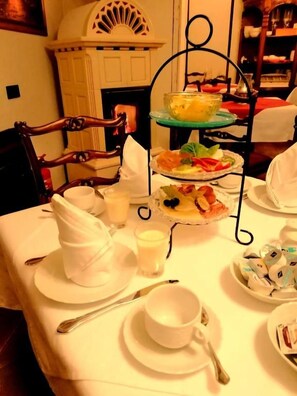 Una mesa de comedor con un soporte de postres de tres niveles, vajilla blanca y una chimenea al fondo.