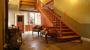 Una escalera de madera con barandilla que conduce a un piso superior. Se ven sillas y una mesa en primer plano, y una zona de bar visible a través de una puerta abierta.