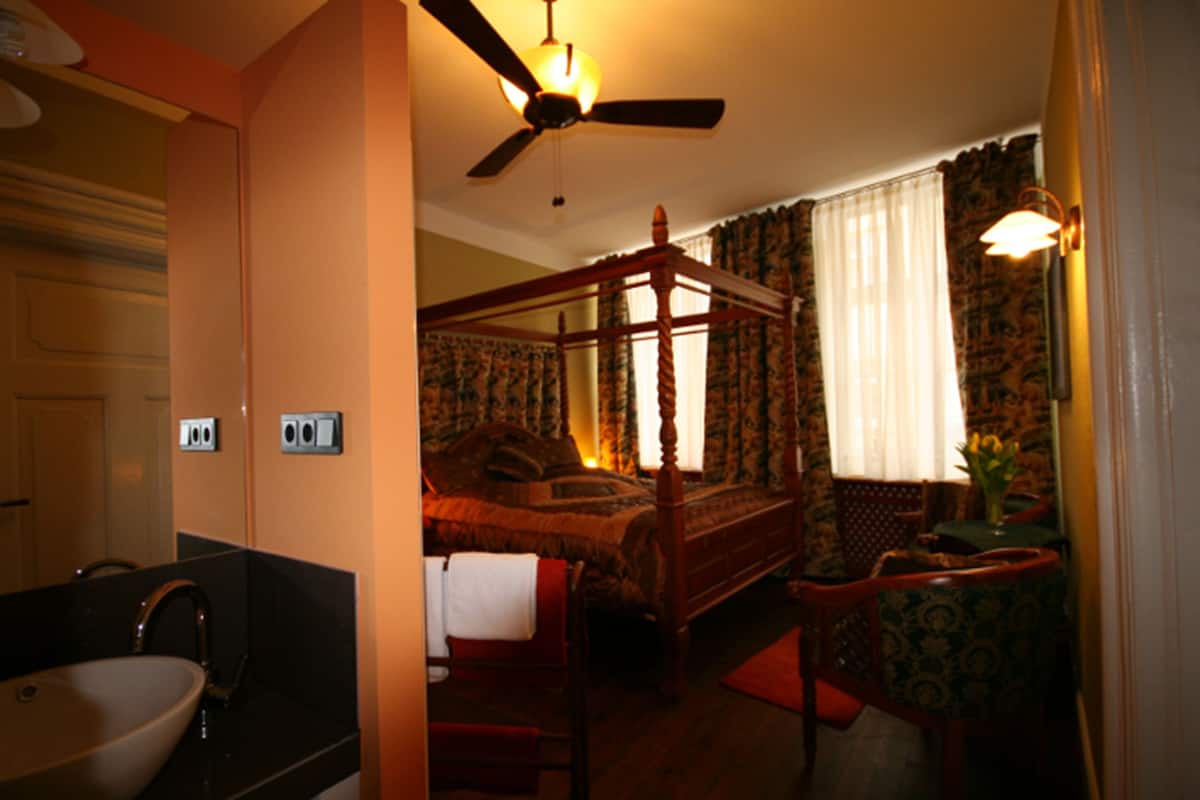 Ein Bett mit Baldachin, ein Holzstuhl, ein kleiner Tisch mit einer Vase und ein Deckenventilator.