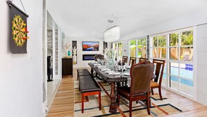 Un espace repas avec une longue table dressée pour six personnes, entourée de chaises et d’un banc en bois, et offrant une vue sur une piscine à travers de grandes fenêtres.