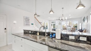Une cuisine moderne avec des meubles blancs, un plan de travail en marbre noir et des appareils électroménagers en acier inoxydable. On y trouve des luminaires suspendus, un escalier menant à un autre niveau et une vue sur l’extérieur grâce à de grandes fenêtres.