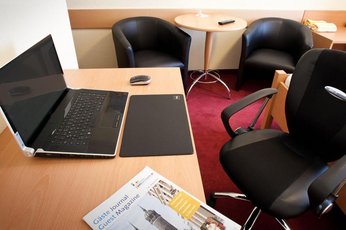 A hotel room with a wooden desk, a laptop, a tablet, a magazine, and two black chairs.