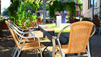 An outdoor seating area with wicker chairs and tables, surrounded by greenery and a building in the background.