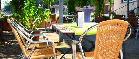 An outdoor seating area with wicker chairs and tables, surrounded by greenery and a building in the background.