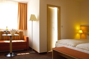 A hotel room with a bed, a sofa, a small table with a lamp, and a window with curtains.