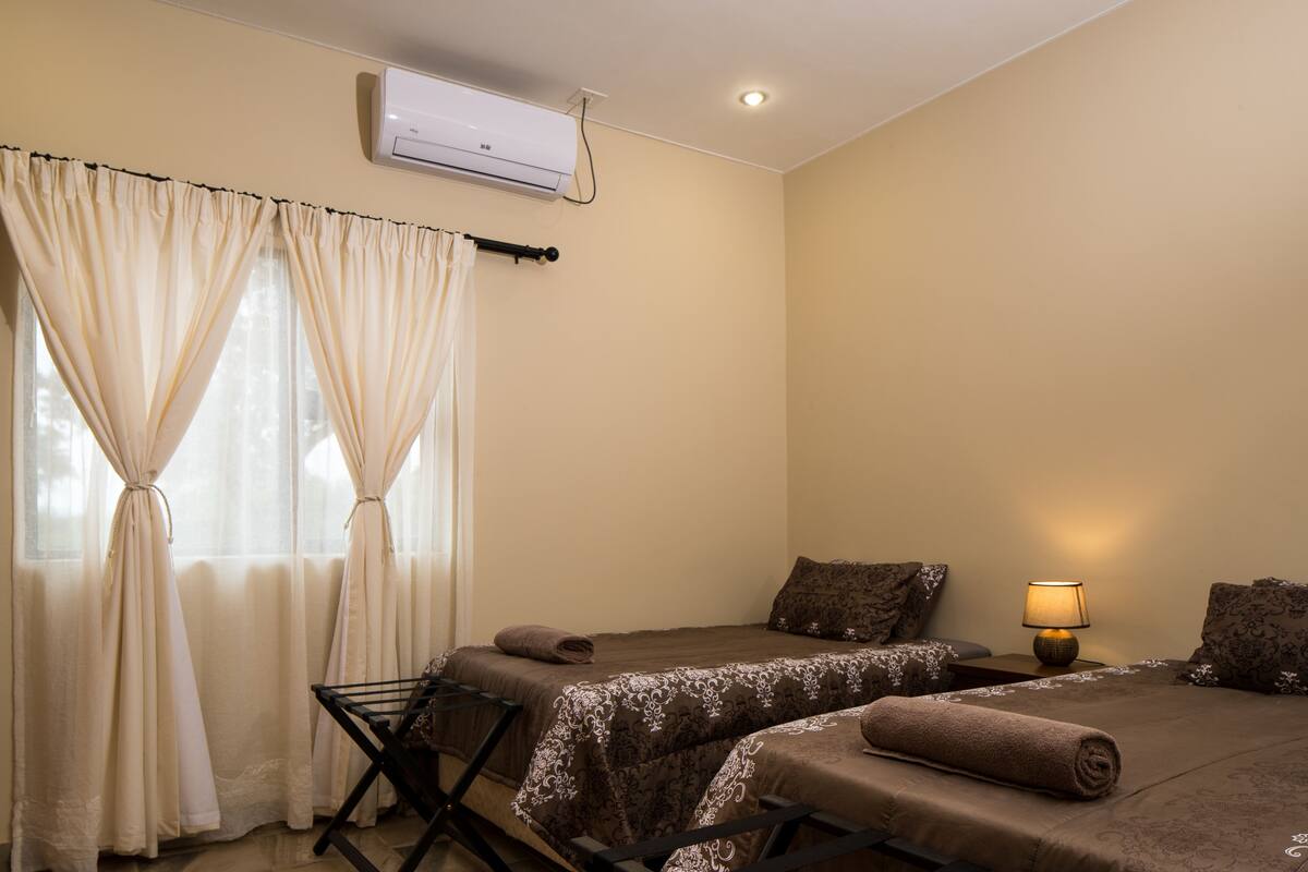 A hotel room with two beds, a ceiling fan, an air conditioning unit, and a bedside lamp.