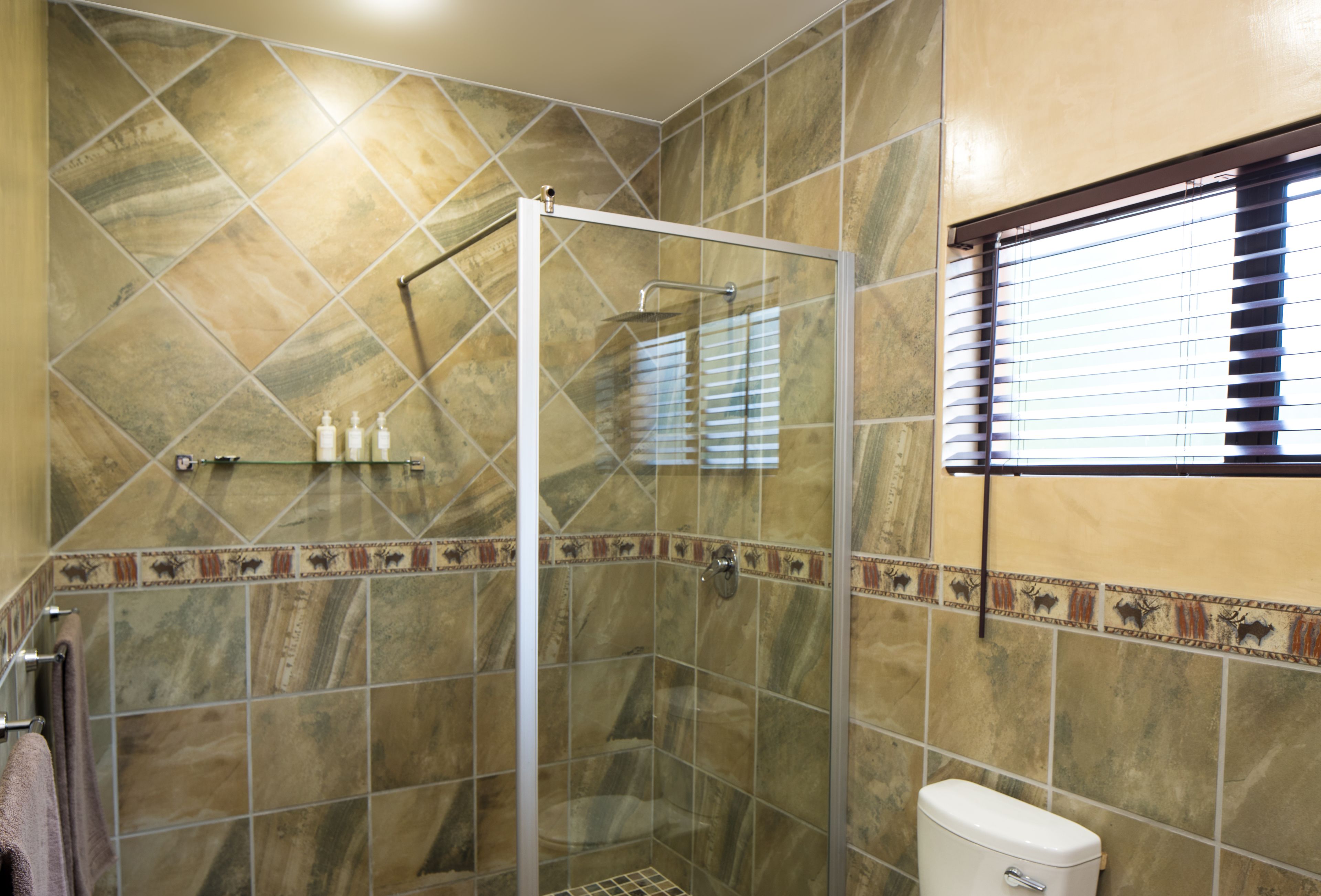 Ein Badezimmer mit einer gläsernen Duschkabine, gefliesten Wänden, einer Toilette und einem Fenster mit Jalousien.
