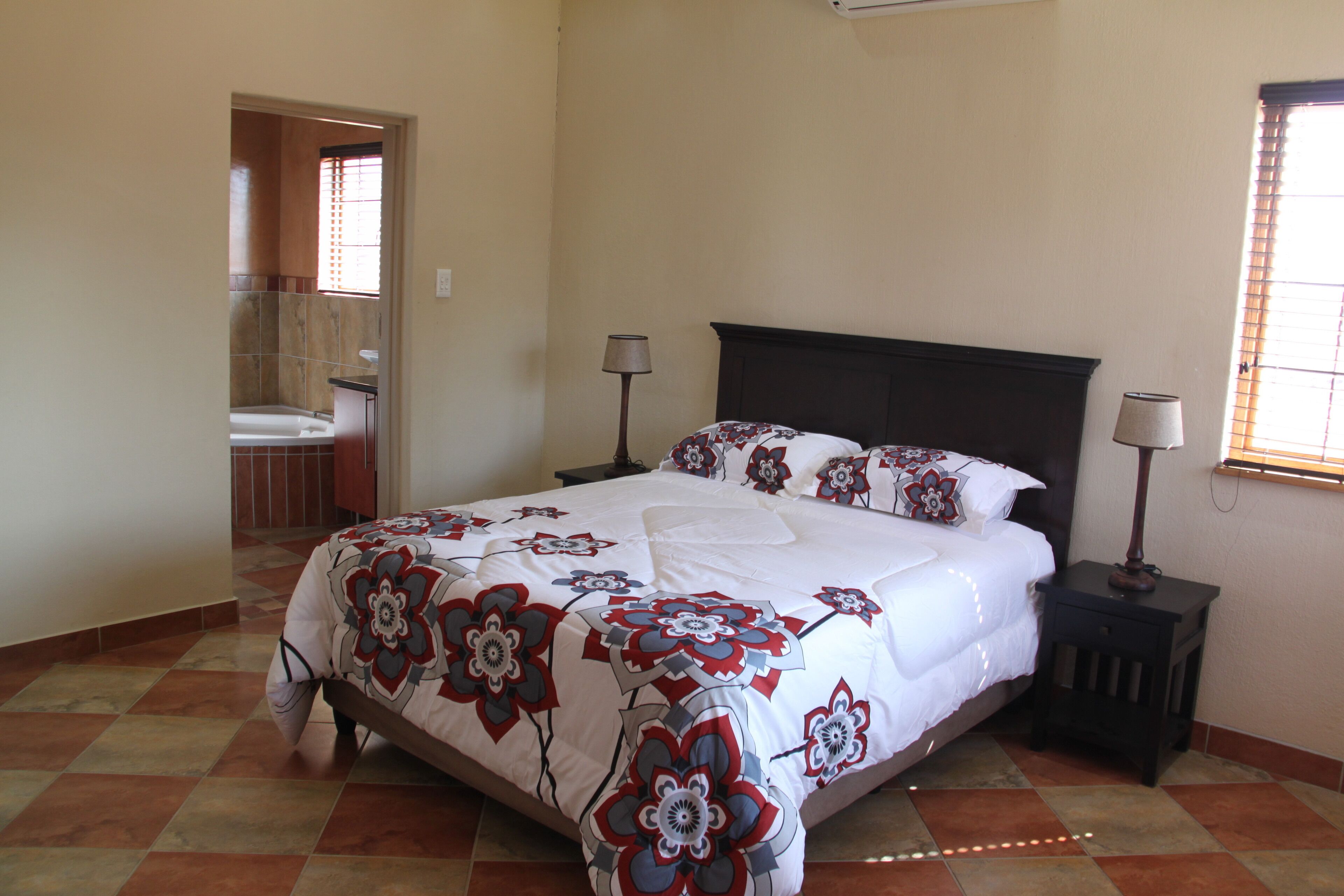 A bedroom with a large bed, two bedside tables with lamps, a bathroom visible through an open door, and a tiled floor.