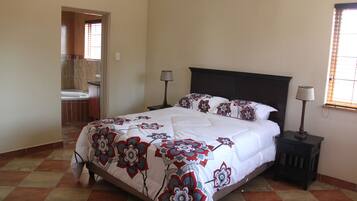 A bedroom with a large bed, two bedside tables with lamps, a bathroom visible through an open door, and a tiled floor.
