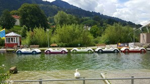 Une rangée de petits bateaux colorés sur une étendue d’eau calme, avec un paysage montagneux en arrière-plan et un petit bâtiment avec un drapeau sur la gauche.