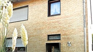 Un bâtiment à deux étages avec une façade en briques, une porte noire et une petite fenêtre. On y trouve des plantes en pot et une zone pavée devant l’entrée.