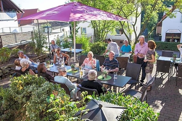 An outdoor patio area with tables and chairs, surrounded by greenery and buildings in the background.