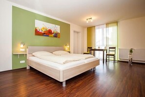A bedroom with a bed, bedside lamps, a painting on the wall, a dining area with a table and chairs, and a window with blinds.