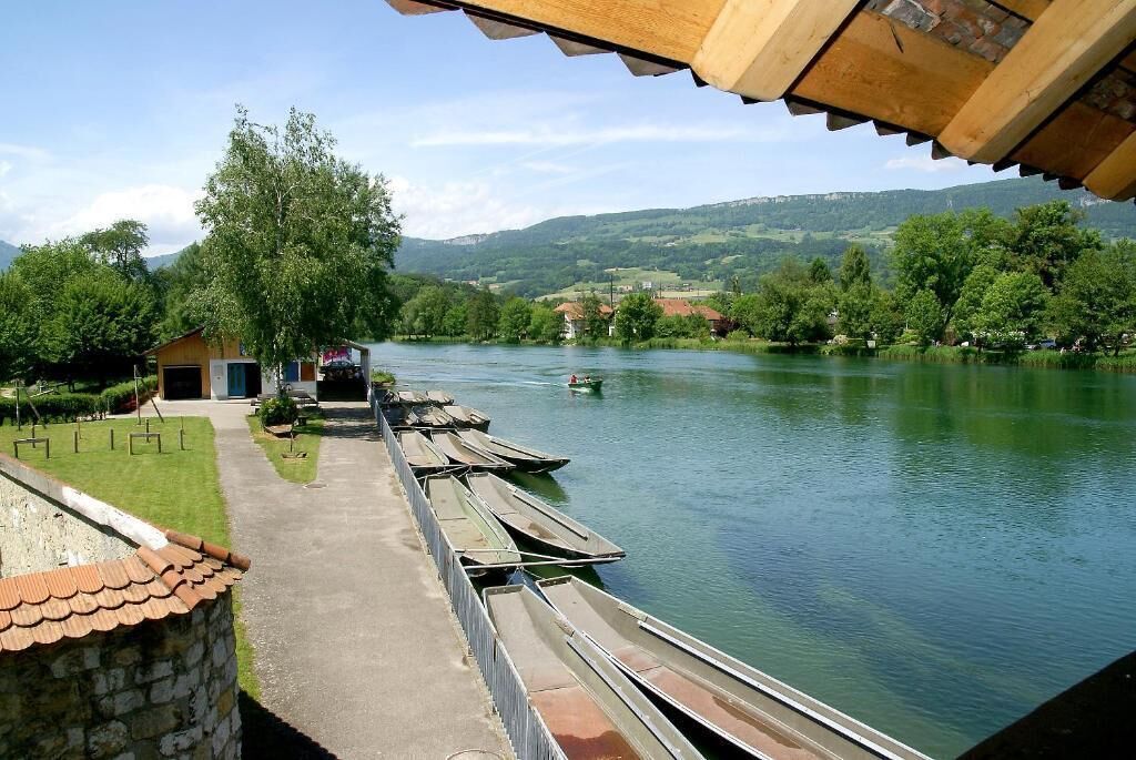 Un quai au bord de la rivière, avec des bateaux amarrés, un petit bâtiment et une vue sur un paysage verdoyant et vallonné.