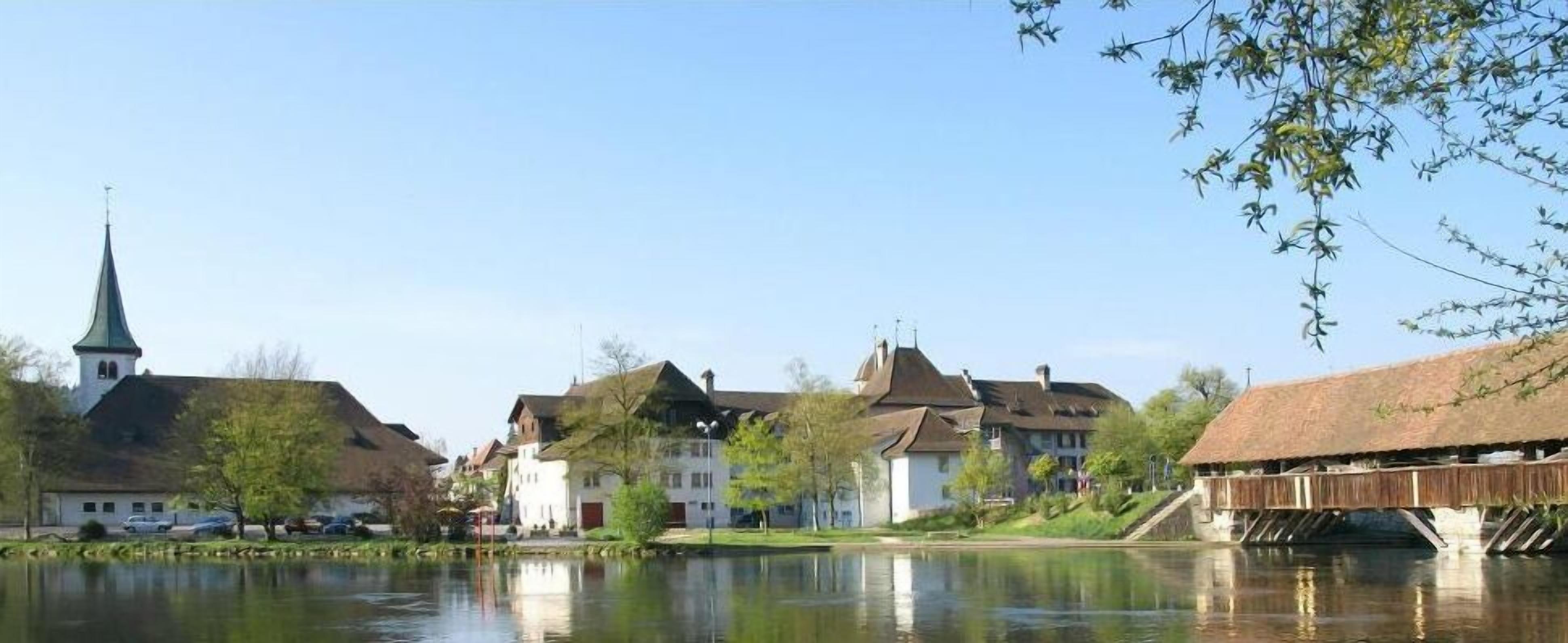 Un pueblo a orillas del río con edificios tradicionales, una aguja de iglesia y un puente de madera.