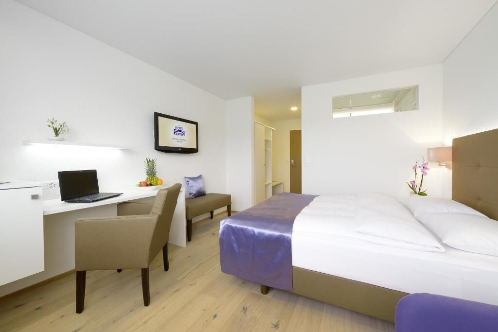 A hotel room with a bed, a desk with a laptop, a chair, a small table, and a wall-mounted shelf with a framed certificate.