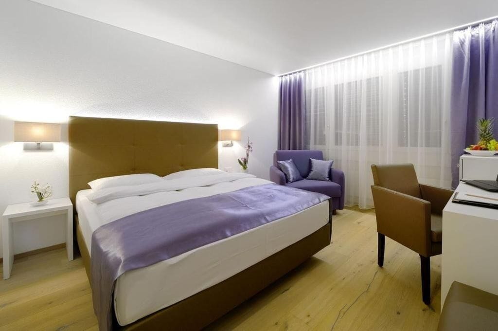 A modern hotel room with a large bed, a desk, a chair, and a window with sheer curtains.