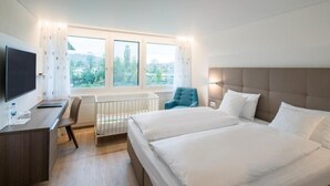 A hotel room with two beds, a desk, a chair, a TV, and a window with a view of greenery. - Hotel Al Ponte (Wiedlisbach)