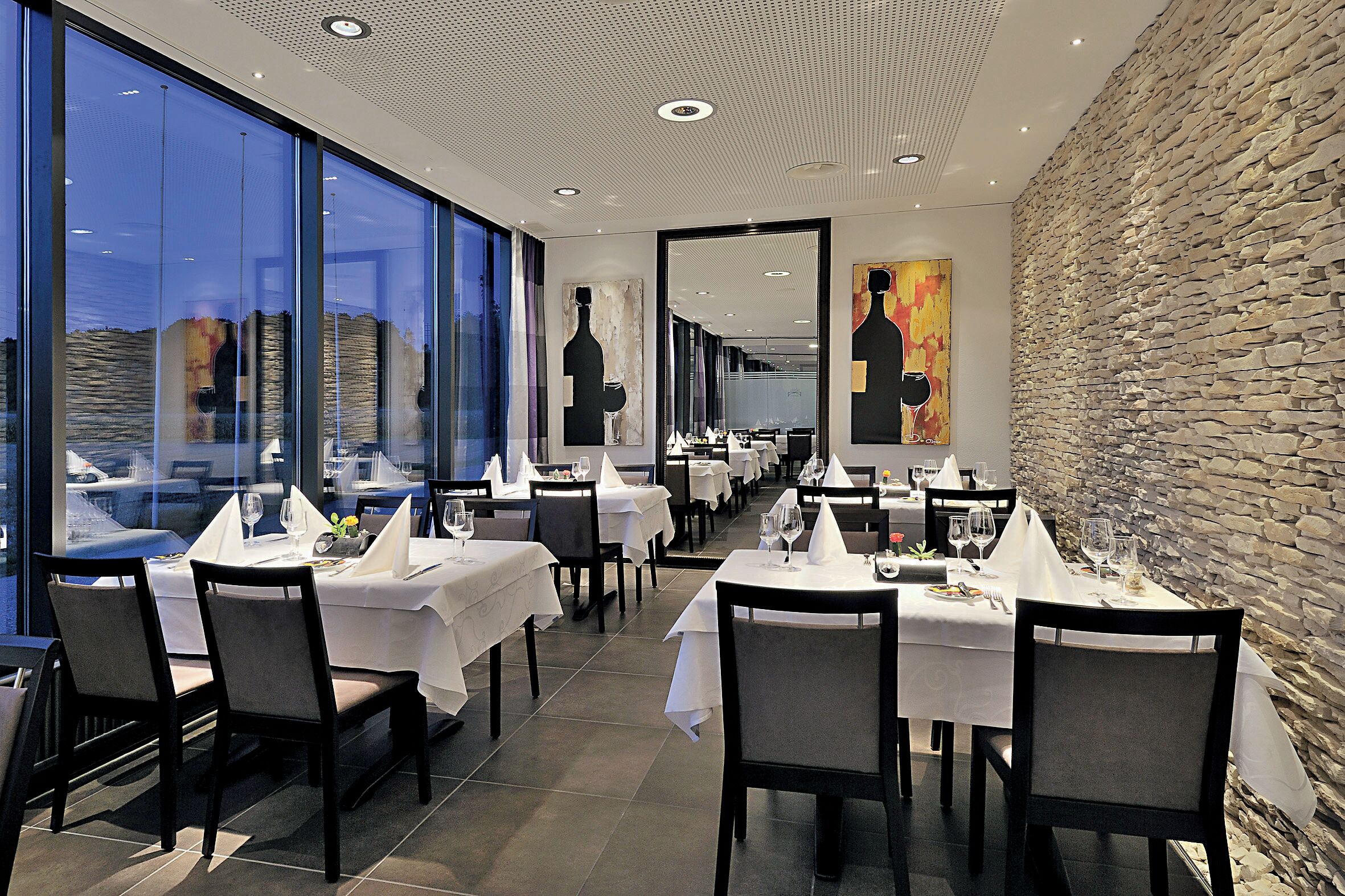 En restaurant med dækkede borde, store vinduer og en stenvæg.