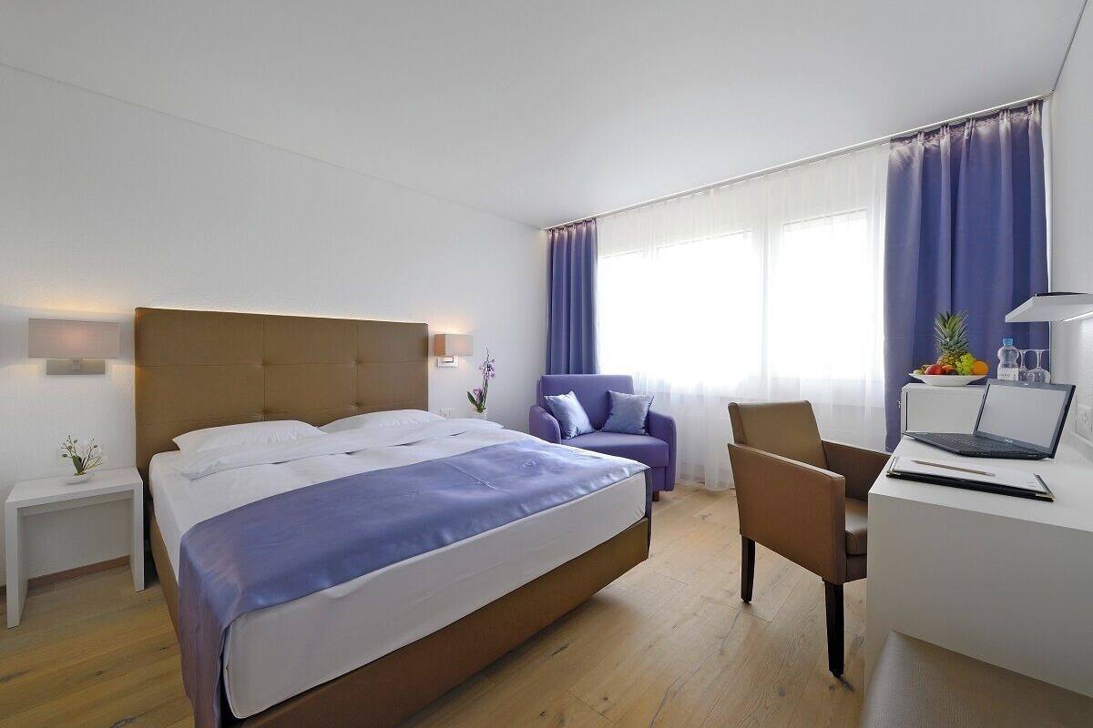 Ein modernes Hotelzimmer mit einem großen Bett, einem Schreibtisch mit einem Laptop, einem Stuhl und einem kleinen Tisch mit Blumen in einer Vase.
