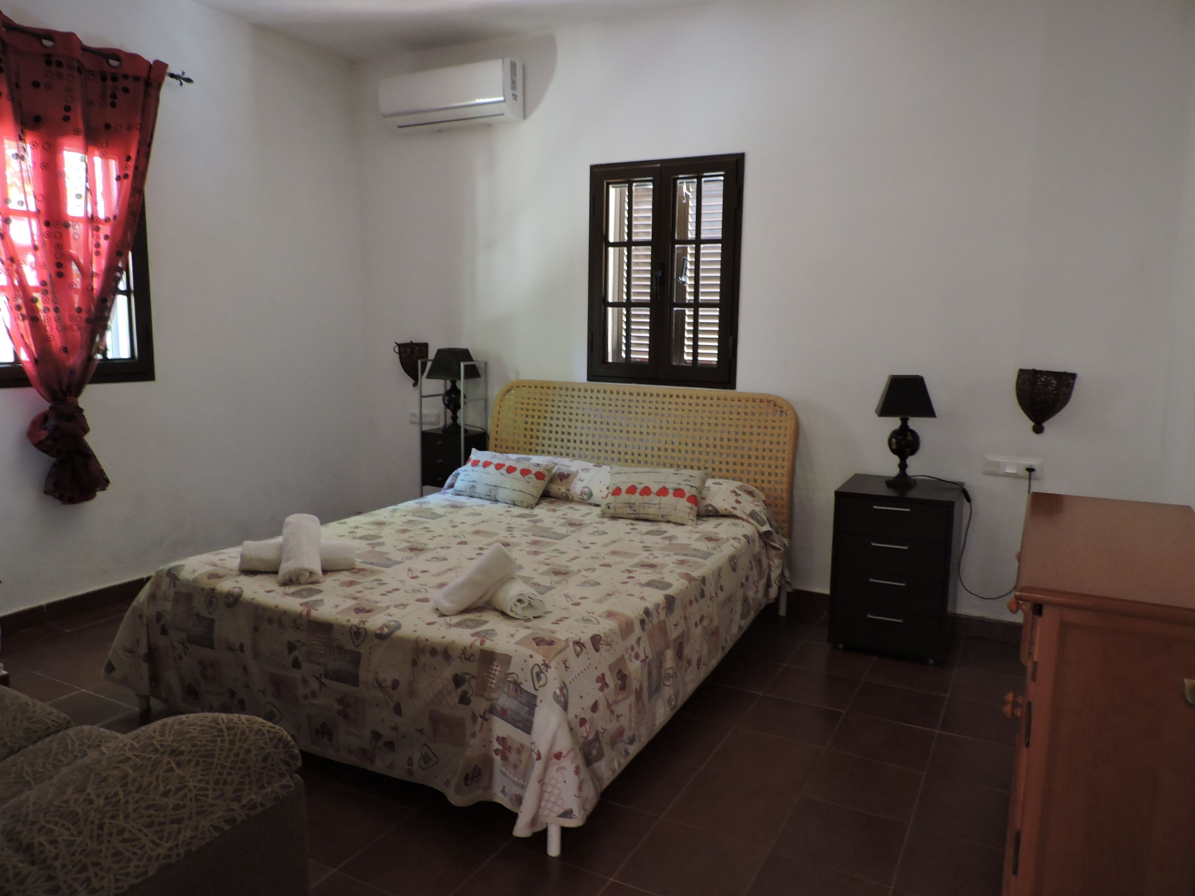 Un dormitorio con cama, mesita de noche, cómoda, ventana con contraventanas, unidad de aire acondicionado y lámpara.