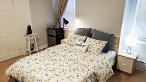 A bedroom with a floral bedspread, a bedside table, a lamp, and a window with curtains. - Lovely, cozy Murray guest suite in Squirrel Hill  (Pittsburgh)
