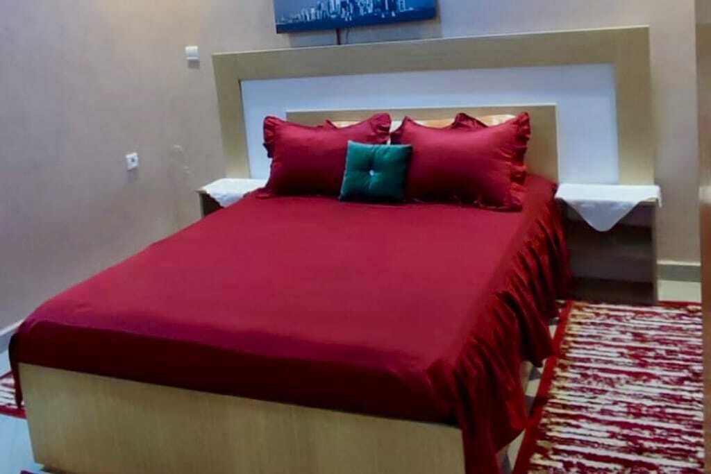 Un lit bien fait, avec une literie rouge et un oreiller vert, une table de chevet blanche et un tapis à motifs rouges et blancs.