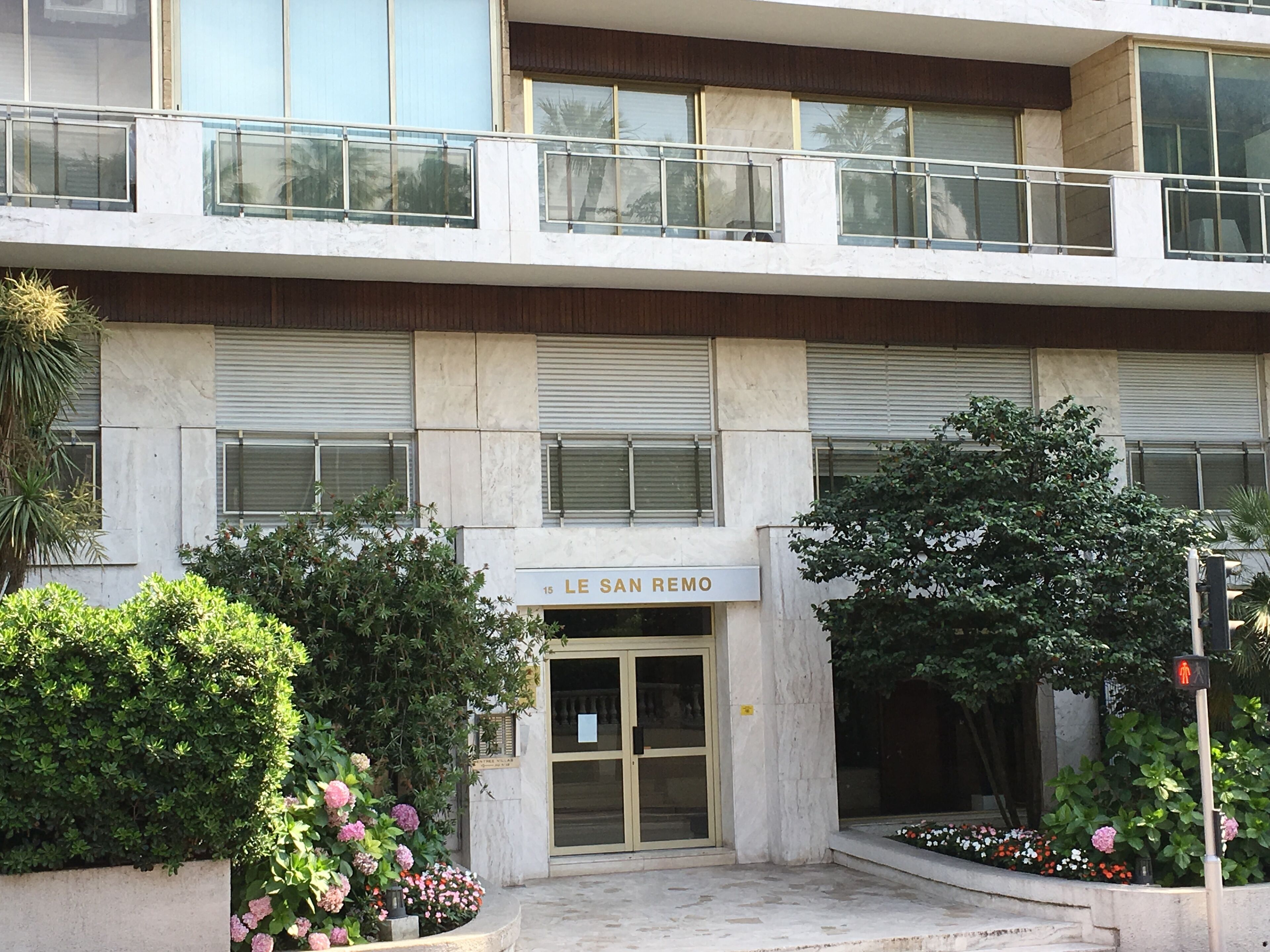A building with a sign reading "LE SAN REMO" at the entrance, featuring a modern design with a glass door, a balcony, and surrounding greenery.