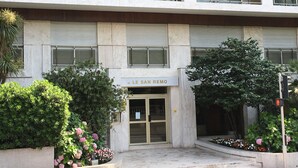A building with a sign reading "LE SAN REMO" at the entrance, featuring a modern design with a glass door, a balcony, and surrounding greenery.