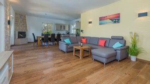 Ein modernes Wohnzimmer mit grauem Ecksofa, Holzboden, Kamin, Essbereich und einer Küche im Hintergrund.