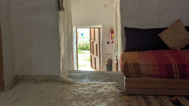 A room with a high ceiling, a window, a bed with a patterned blanket, a small table with a teapot, and a wall-mounted radio.