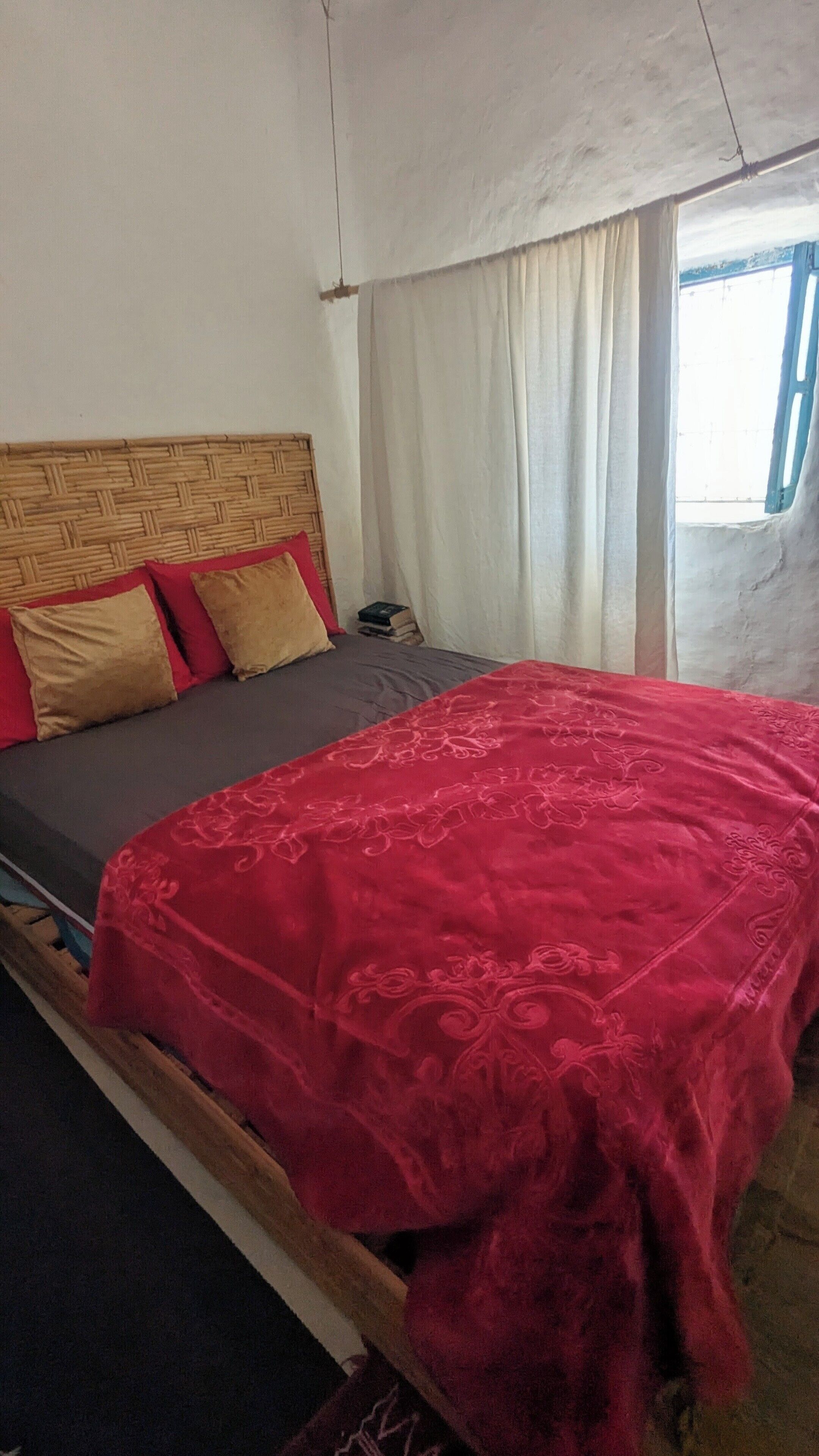 A single bed with a red bedspread, a wooden headboard, and a window with curtains.