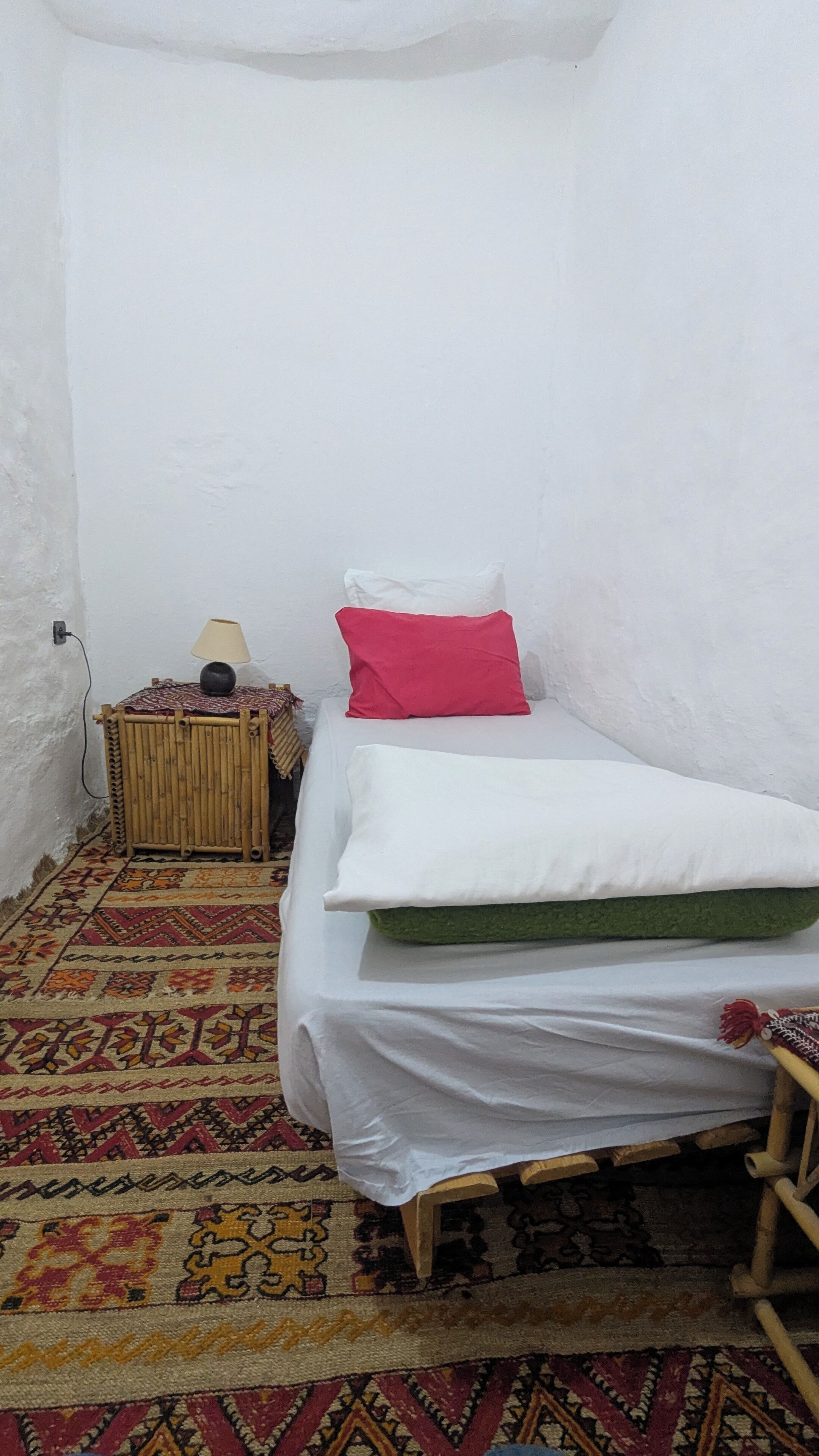 A small, single-bed room with a patterned rug, a bedside table with a lamp, and a red pillow.