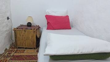 A small, single-bed room with a patterned rug, a bedside table with a lamp, and a red pillow.