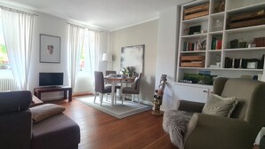 Ein Wohnzimmer mit Sofa, Couchtisch, Fernseher, Esstisch mit Stühlen und Bücherregal.