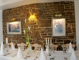 Una mesa de comedor dispuesta para una comida, con mantel blanco, velas y cuadros en una pared de piedra.