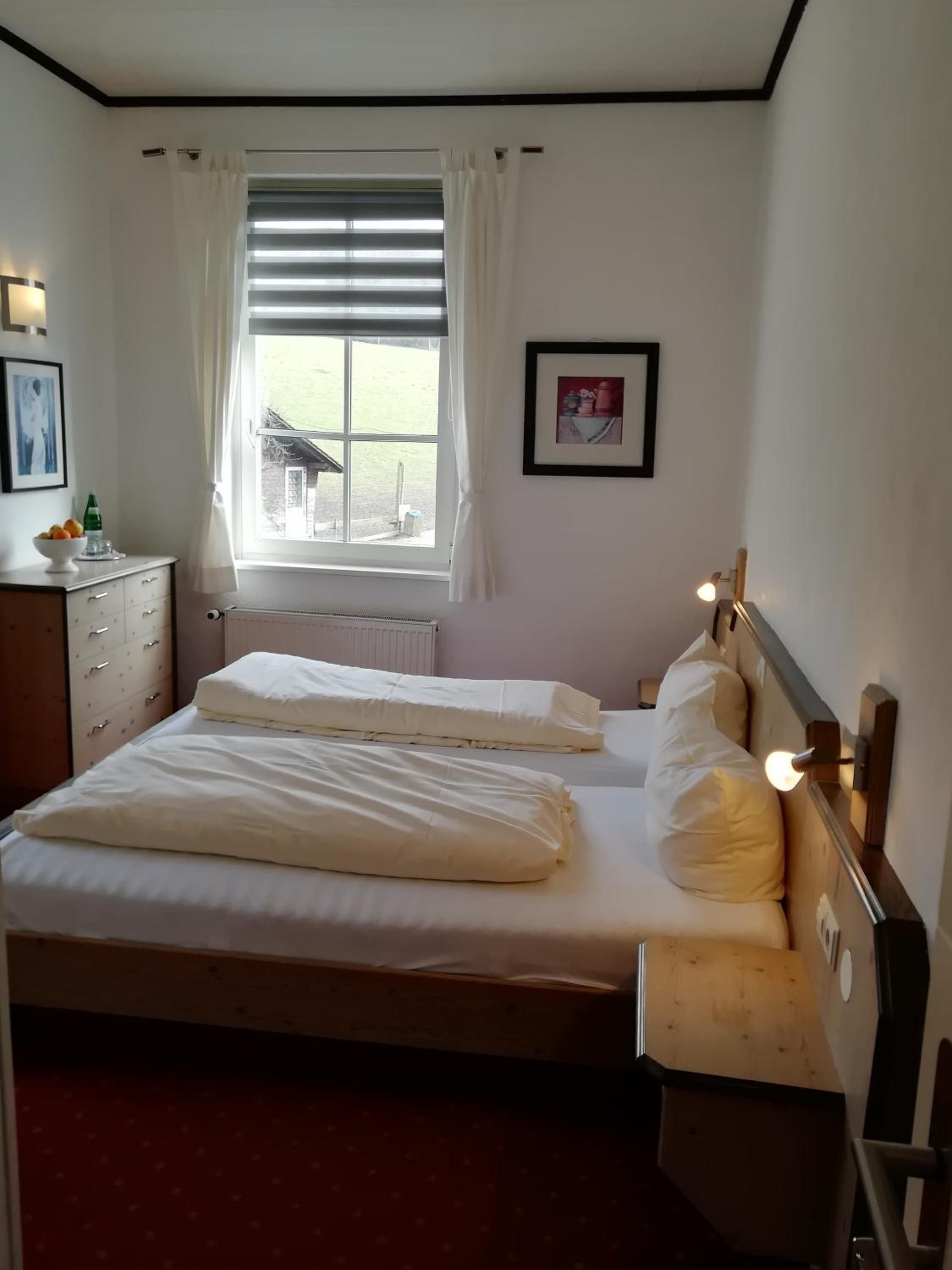 A bedroom with two beds, a window with blinds, a radiator, a bedside table with a lamp, and a framed picture on the wall.