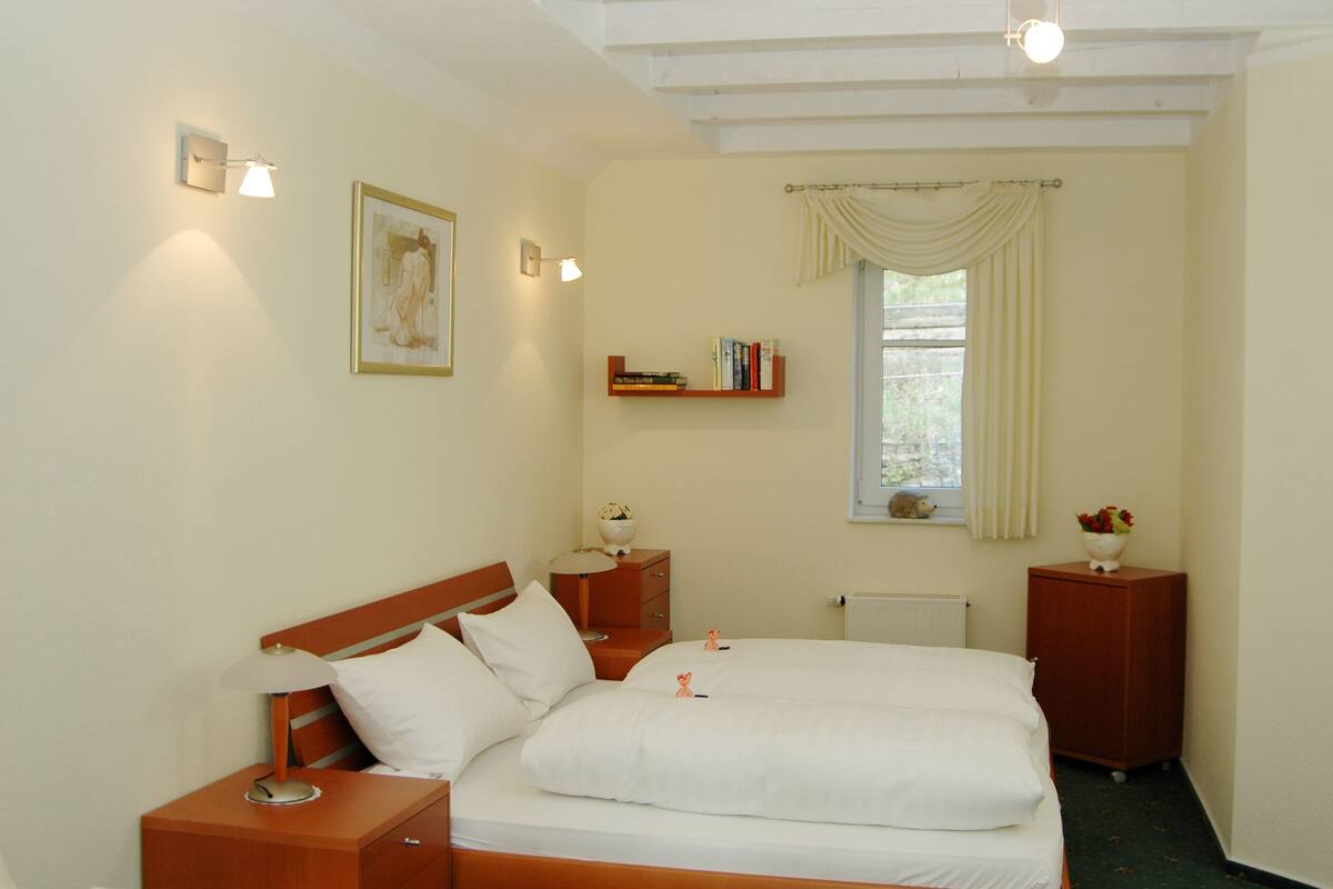 A neatly arranged bedroom with a bed, bedside tables, a shelf with books, a window with curtains, and a radiator.