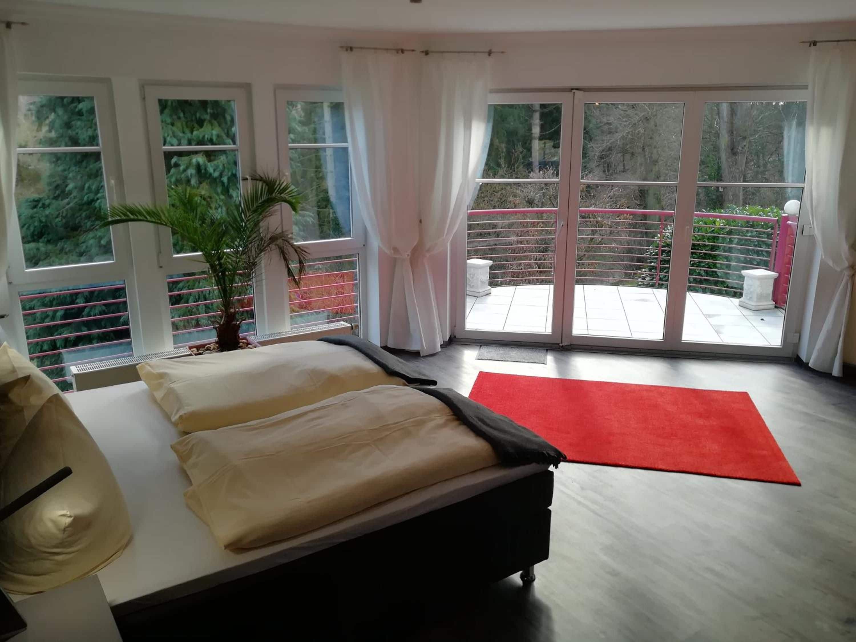 Un dormitorio con cama, alfombra roja, planta en maceta y vista a un balcón con barandilla.