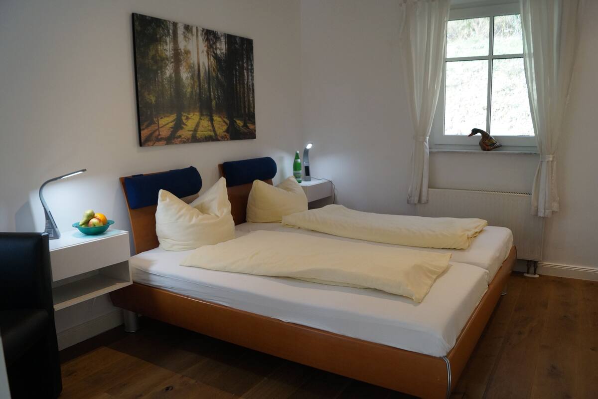 A double bed with white bedding, a nightstand with a lamp and a bottle, a desk with a chair and a bowl of fruit, a painting of a forest, and a window with a view of trees.
