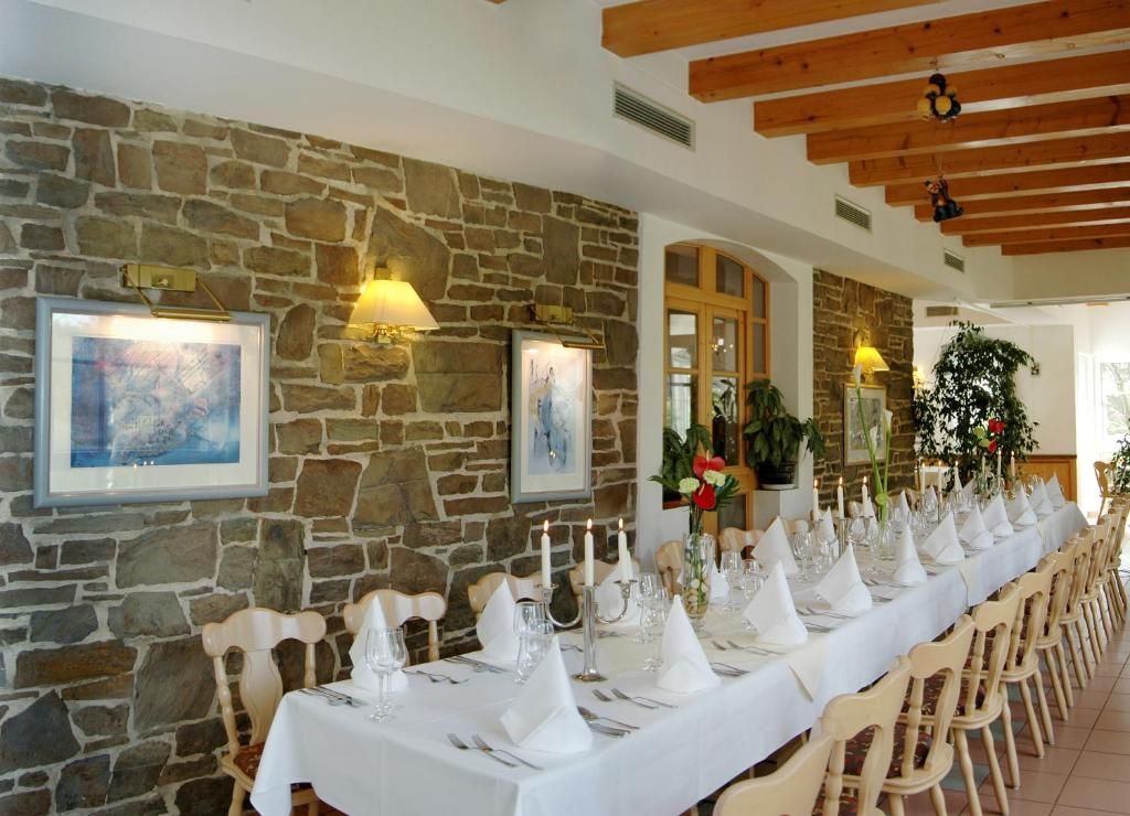 Una larga mesa de comedor preparada para una comida, con manteles y servilletas blancos, sillas de madera y cuadros en la pared de piedra.