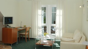 A room with a desk, chair, and a sofa. There is a window with curtains, a television on a cabinet, and a table with a vase of flowers. - Waldhotel Forsthaus Remstecken (Koblenz)
