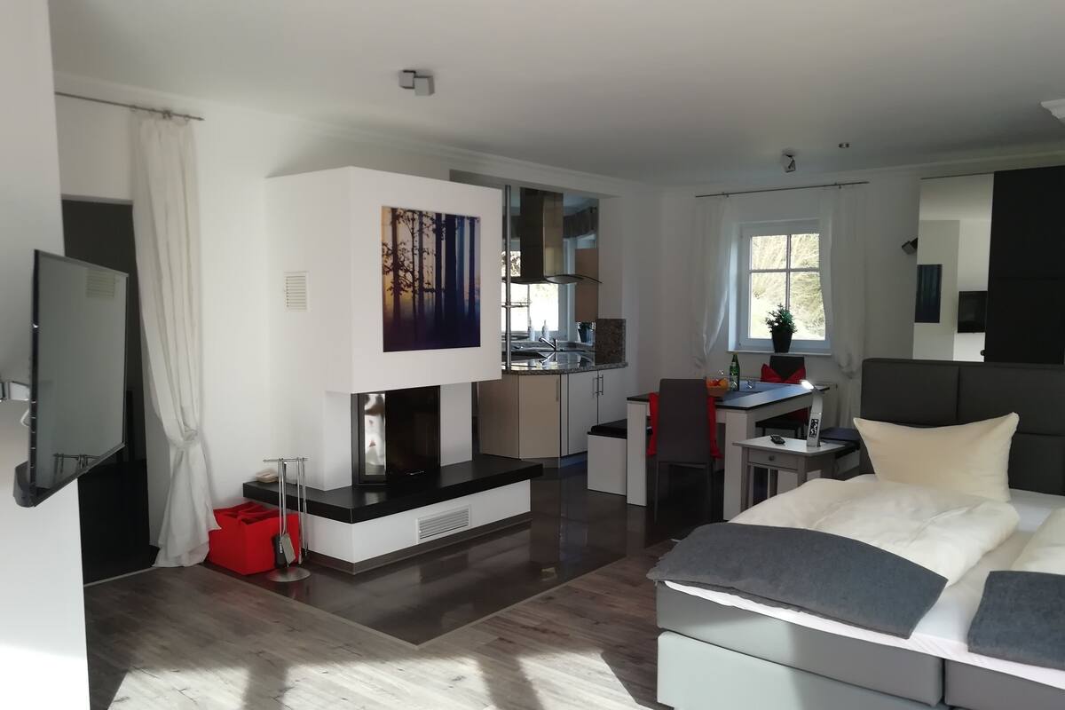 A modern hotel room with a large bed, a desk, a kitchenette, and a TV.